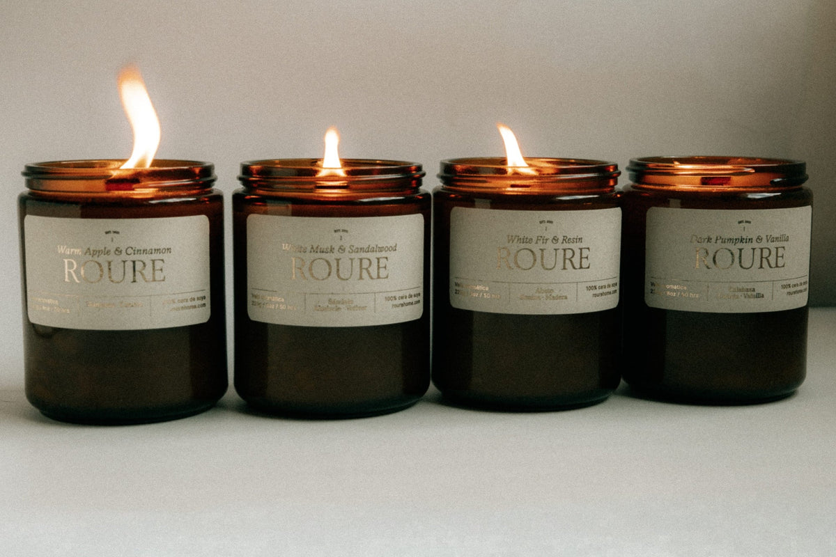 Four lit amber glass jar candles with white labels are lined up against a neutral light gray background. The labels show different scent names including Warm Apple & Cinnamon, Roure, White Tea & Rose, and Dark Pumpkin & Sandalwood.