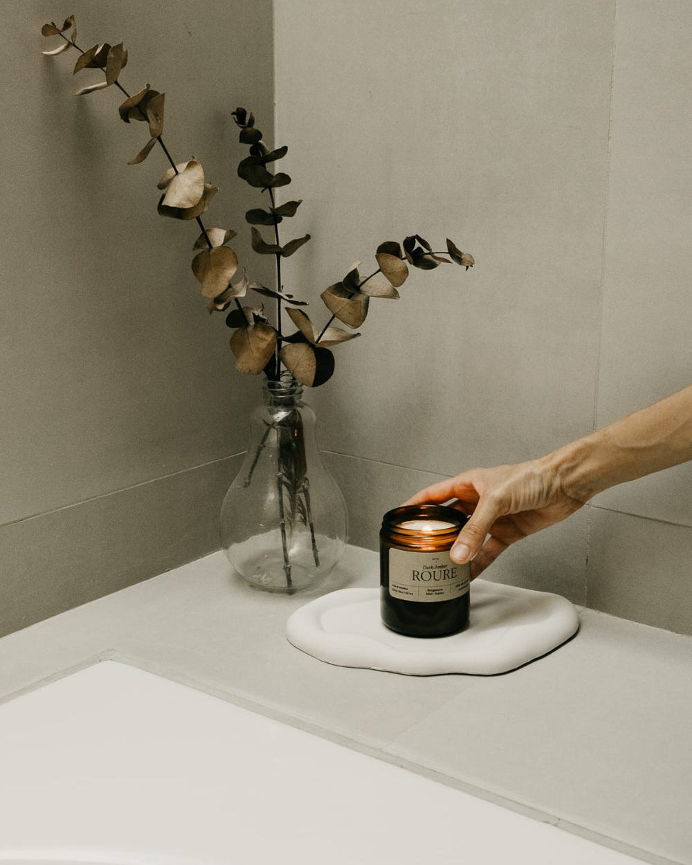 Hand holding a candle next to a vase with dried flowers on a neutral background