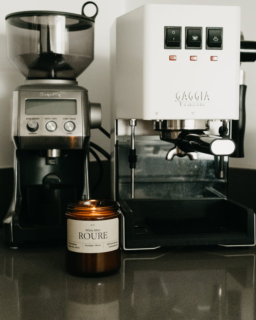 A countertop setup featuring a silver and black coffee grinder next to a white La Moka espresso machine, with a brown amber glass candle labeled 'Rouge' in front.