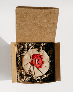 A round beige item wrapped in paper and sealed with a red wax seal featuring a monogram letter 'R', presented inside an open brown kraft gift box with crinkle paper filler on a white background.