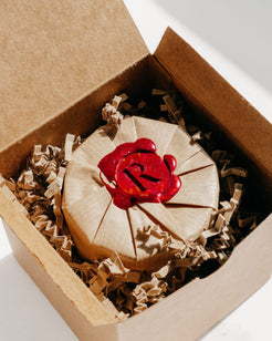 A round kraft paper-wrapped item sealed with a red wax seal featuring a stylized letter 'R', presented inside an open brown cardboard box with crinkle paper filler.
