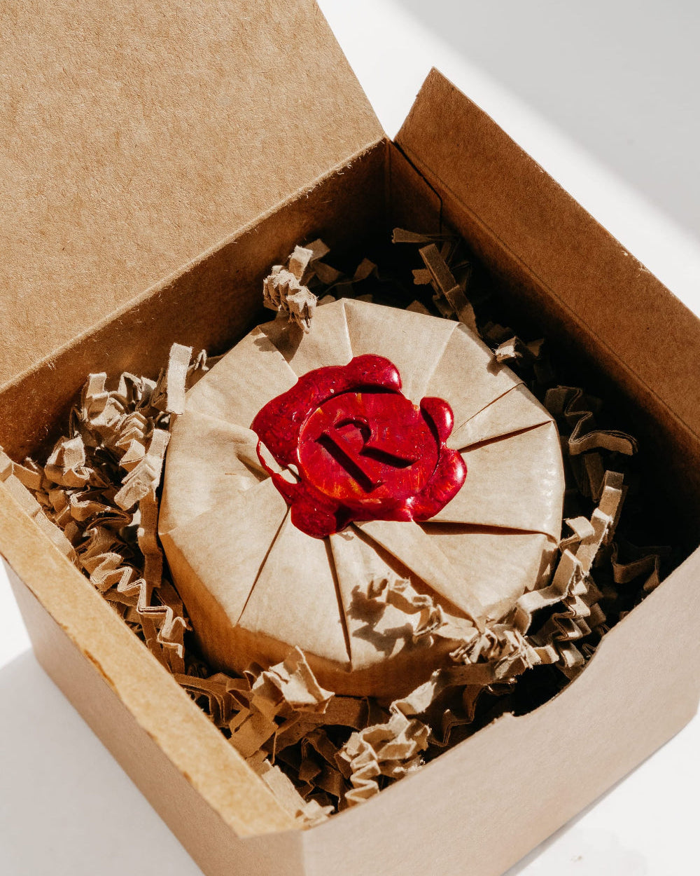 A round kraft paper-wrapped item sealed with a red wax seal featuring a stylized letter 'R', presented inside an open brown cardboard box with crinkle paper filler.
