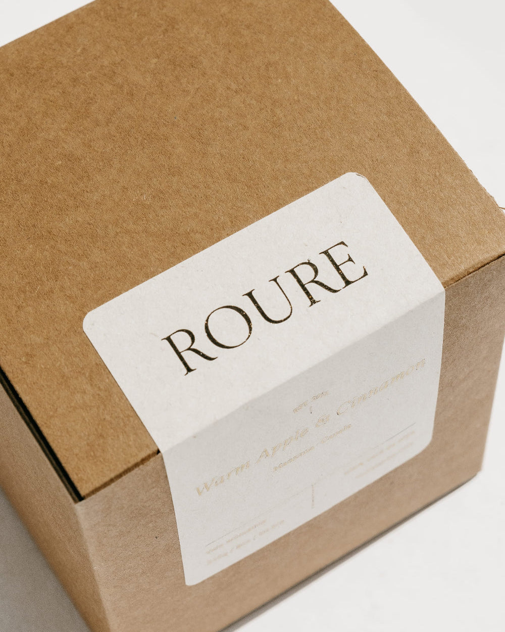 A square kraft paper gift box with a white paper label that reads 'ROURE' in dark serif lettering, photographed on a clean white background.