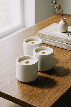 Three white candles in ceramic holders on a wooden surface with books and a vase in the background.